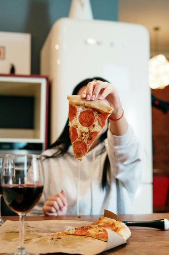 Person hält ein Stück Pizza hoch, im Hintergrund unscharf ein Weinglas und eine stilvolle Küche