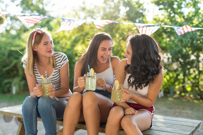 Drei lachende Teenagerinnen trinken erfrischende alkoholfreie Limonade bei einer Gartenparty, ein Bild, das den Spaß jugendlicher Zusammenkünfte ohne Alkohol darstellt