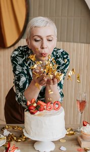  Frau pustet Konfetti von den Händen, feiert den 60. Geburtstag mit Kuchen und Sektgläsern im Hintergrund