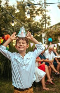 in glücklicher Junge feiert im Garten bei Tageslicht seinen Geburtstag, lacht und balanciert einen Partyhut auf seinem Kopf, während im Hintergrund Erwachsene an einem festlich dekorierten Tisch sitzen und den Moment genießen.