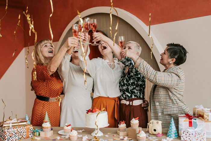 Gruppe fröhlicher Menschen stößt mit "alkoholfreiem Sekt"* an, um einen Geburtstag zu feiern, umgeben von festlicher Dekoration und süßen Leckereien