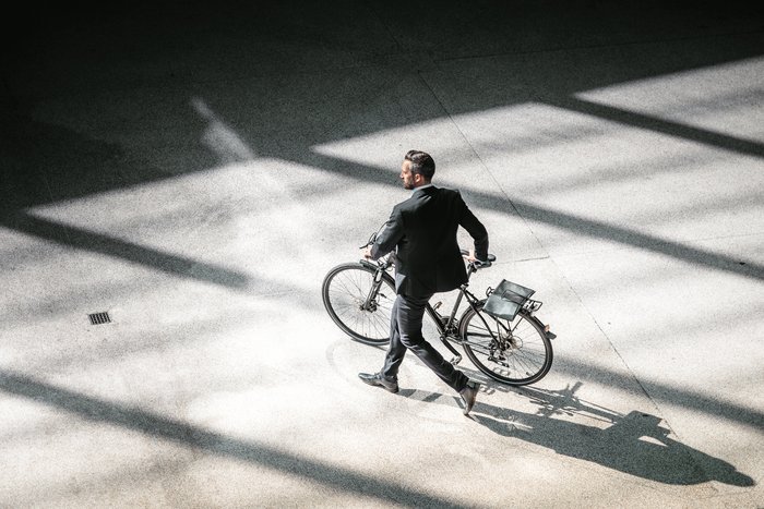 Geschäftsmann schiebt sein Fahrrad über einen sonnenbeschienenen Platz, was das Bewusstsein für die Bedeutung der Nüchternheit beim Radfahren auf der Seite "Alkohol & Radfahren" betont.