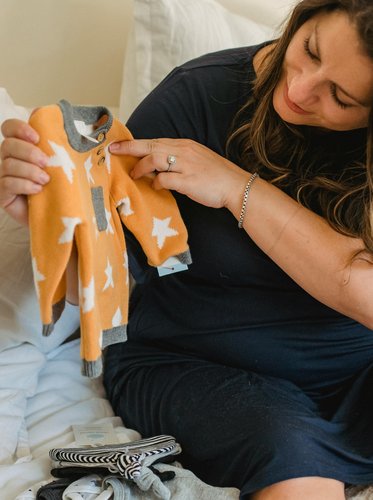 Schwangere Frau sitzt auf Bett und betrachtet liebevoll ein kleines orange-weißes Babyjäckchen