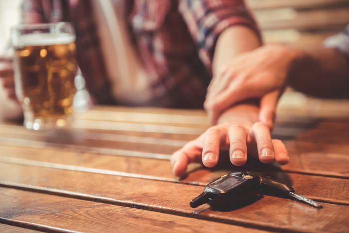 Hand einer Person, die neben einem Glas Bier und Autoschlüsseln auf einem Holztisch liegt, symbolisiert die Wichtigkeit der Entscheidung gegen Alkohol am Steuer auf der Informationsseite zum Thema "Alkohol am Steuer"