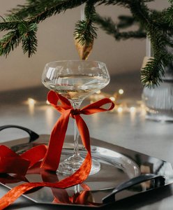 Festlich dekoriertes Weinglas mit rotem Band unter Tannenzweigen, stimmungsvolle Weihnachtsbeleuchtung im Hintergrund
