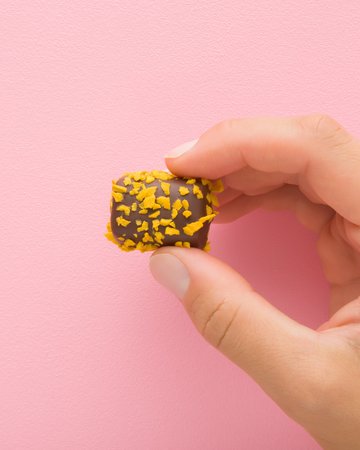 Eine Hand hält eine Praline vor einer rosa Wand, die mit goldgelben Splittern bestreut ist